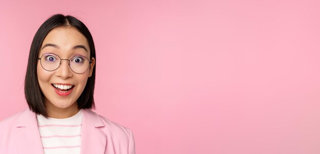 Крупный план портрета азиатской деловой женщины в очках, удивленной реакцией камеры, стоящей в костюме на розовом фоне
