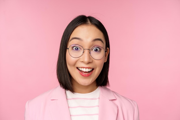 Close up portrait of asian businesswoman in glasses looking surprised at camera amazed reaction standing in suit over pink background