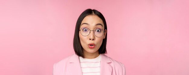 Close up portrait of asian businesswoman in glasses looking surprised at camera amazed reaction standing in suit over pink background