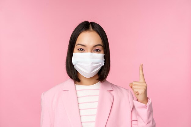 Close up portrait of asian businesswoman in medical face mask and suit pointing finger up showing advertisement top banner standing over pink background