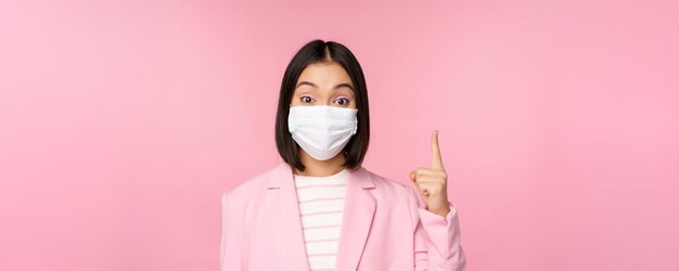 Крупный план портрета азиатской деловой женщины в медицинской маске и костюме, указывающей пальцем вверх, показывая рекламный верхний баннер, стоящий на розовом фоне