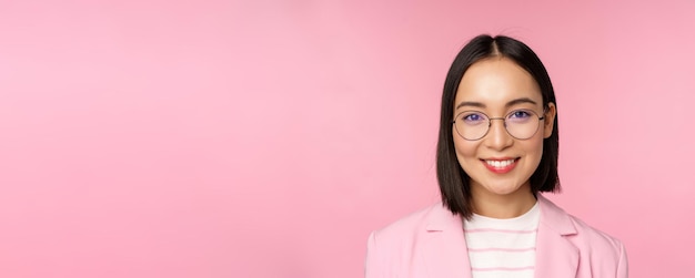 Free photo close up portrait of asian corporate woman professional businesswoman in glasses smiling and looking confident at camera pink background