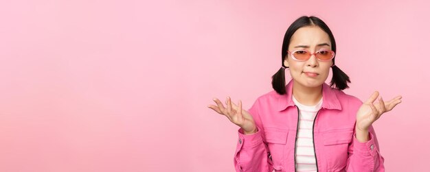 Close up portrait of asian girl looking confused shrugging puzzled and looking at camera wearing sunglasses standing over pink background