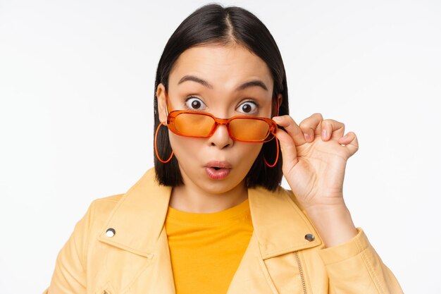 Close up portrait of asian girl looking surprised wow face takes off sunglasses and staring impressed at camera standing over white background