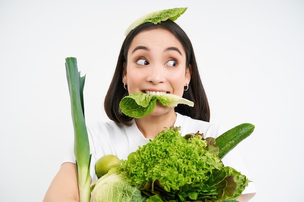 Close up portrait of asian girl munching lettuce leaf looking excited holding pile of vegetables gre