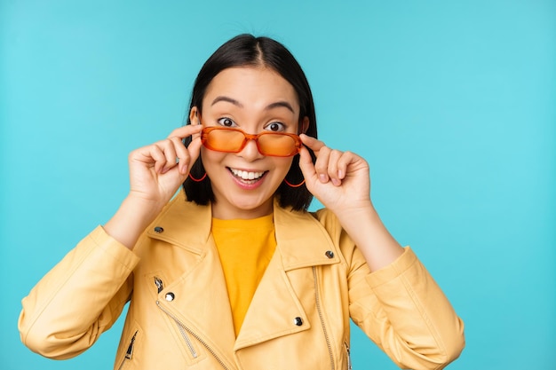 Close up portrait of asian girl in sunglasses looking happy and surprised excitement on face standing over blue background