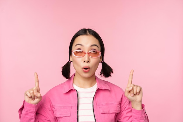 Free photo close up portrait of asian woman gasping looking surprised pointing finger at banner advertisement standing over pink background