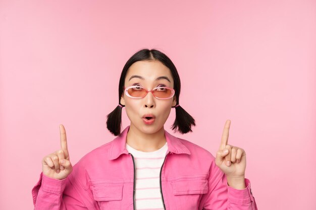 Close up portrait of asian woman gasping looking surprised pointing finger at banner advertisement standing over pink background