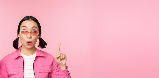 Close up portrait of asian woman gasping looking surprised pointing finger at banner advertisement standing over pink background