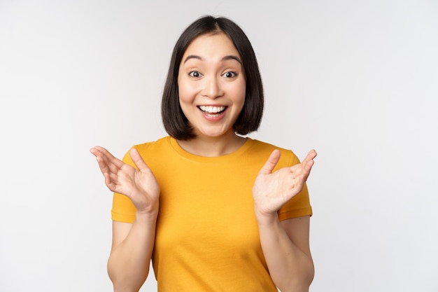 Close up portrait of asian woman looking surprised wow face staring impressed at camera standing over white background in yellow tshirt