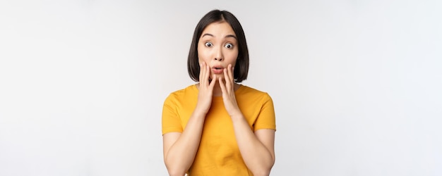 Close up portrait of asian woman looking surprised wow face staring impressed at camera standing over white background in yellow tshirt