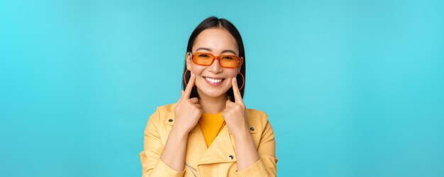 선글라스를 끼고 웃고 있는 아시아 젊은 여성의 초상화를 클로즈업하고 파란 배경 위에 행복해 보이는 낭만적인 서 있는 모습