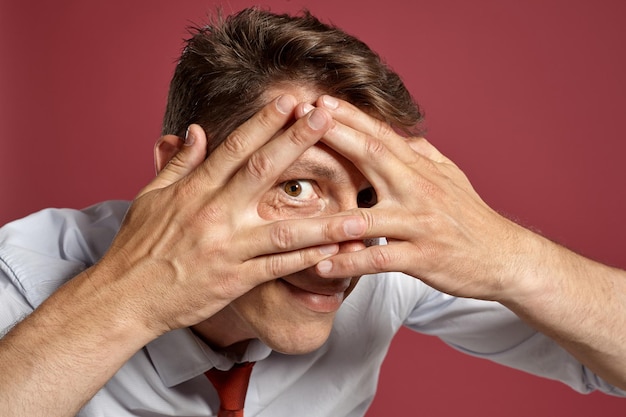 Free photo close-up portrait of an athletic brunet fellow with brown eyes, wearing in a white shirt and a terracotta tie. he is fooling around while posing in a studio against a red background. concept of gestic