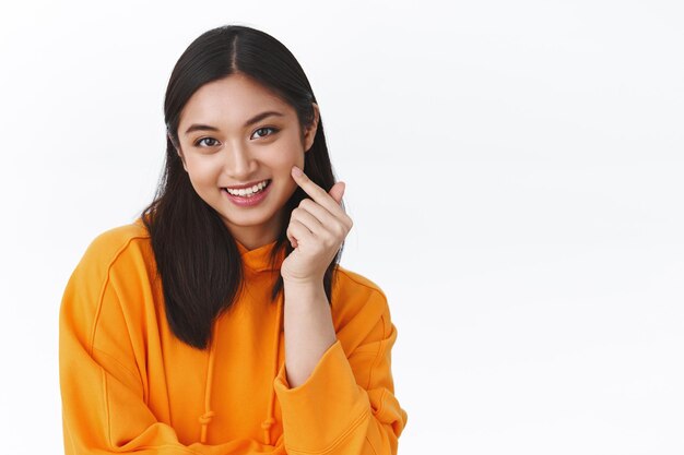 Close-up portrait attractive asian woman in orange hoodie promote skincare products, cosmetology care, pointing face and smiling delighted, got rid of acne