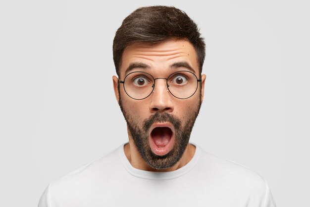 Close up portrait of attractive beared man reacts on sudden news, stares through spectacles, opens mouth widely