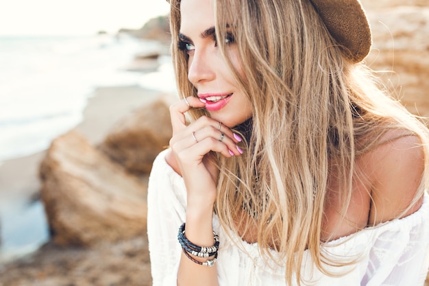 Close-up portrait of attractive blonde girl with long hair on deserted beach. She keeps finger on lips and looks far away.