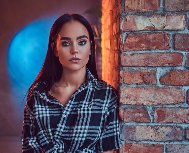 Close-up portrait of an attractive brunette dressed in flannel shirt leaning on the wall in a room with loft interior.