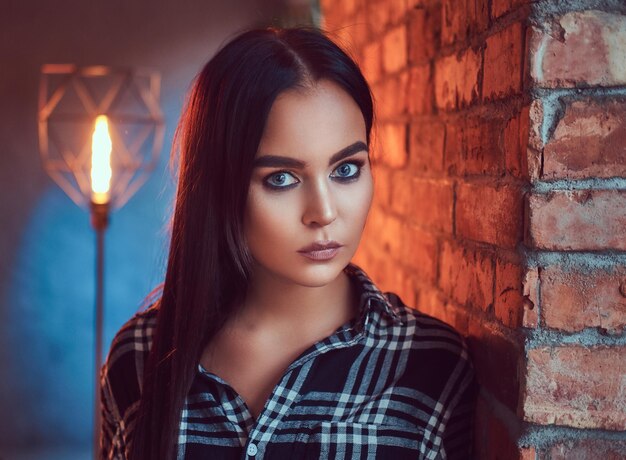 Close-up portrait of an attractive brunette dressed in flannel shirt leaning on the wall in a room with loft interior.