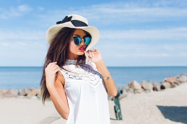 Close-up portrait of attractive brunette girl with long hair standing on the beach near sea. She is touching hair and looking far away.