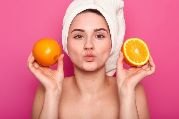 Close up portrait of attractive cheerful woman holds orange slices, keeps lips folded, wears towel and bare shoulders, poses on pink. Model poses in studio. Natural beauty concept.