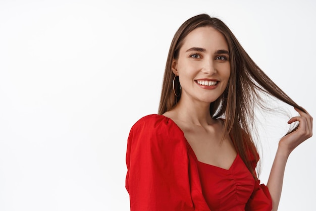 Close up portrait of attractive and confident woman playing with hair strand look flirty and smiling at camera standing in red dress against white background Copy space