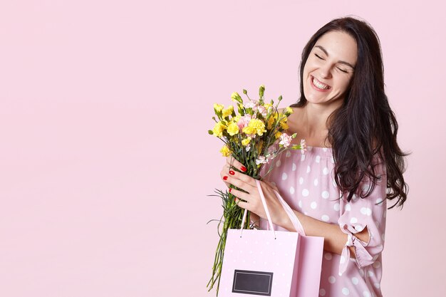 Close up portrait of attractive dark haired young woman in dress, enjoys spring coming, likes to get flowers and presents from her hasband, screw eyes hapily. People, presents, celebration concept.