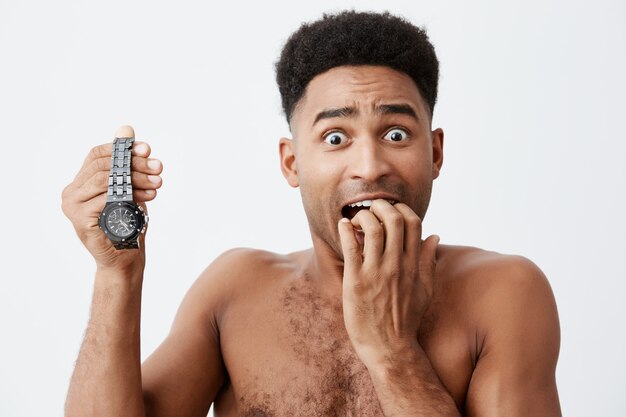 Close up portrait of attractive sexy black skinned american guy with curly hair without clothes holding hand watch in hand, holding hand near mouth, looking in camera with frightened expression.