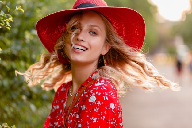 わらの赤い帽子とブラウスの夏のファッションの衣装で笑顔の巻き毛の髪型で魅力的なスタイリッシュな金髪の笑顔の女性のクローズアップの肖像画