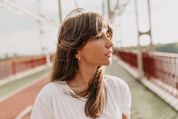 Close up portrait of attractive stylish woman with long wavy hair is looking aside and enjoying sun Outside photo of lovely smiling girl walking on bridge