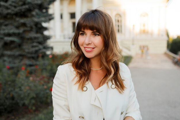 Close up portrait of  attractive  sucsessful business woman posing outdoor. Wavy hairs, white elegant jacket.