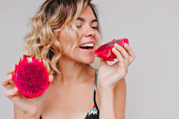 Close-up portrait of attractive tanned woman with short hairstyle eating dragon fruit. refined girl enjoying juicy red pitaya.