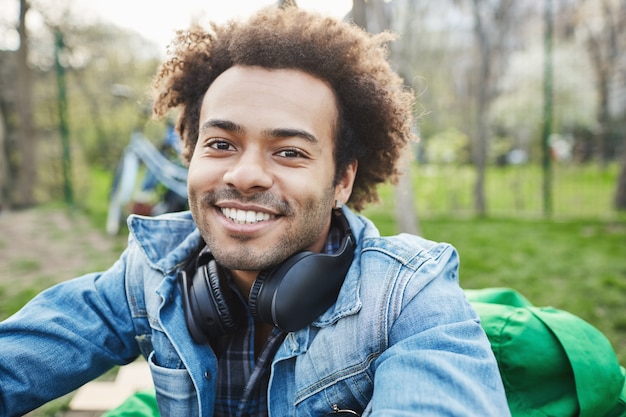 Крупным планом портрет привлекательного небритого темнокожего мужчины с афро-прической, улыбающегося и выражающего счастье, сидя в парке, наслаждаясь хорошей погодой и слушая музыку.