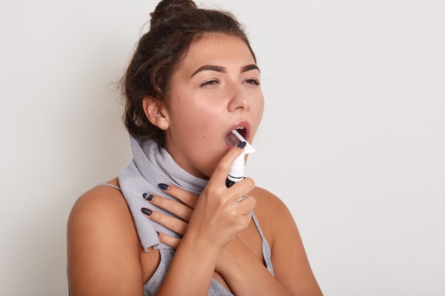 Close up portrait of attractive upset young brunette woman using spray to treat sore throat, painful swallowing