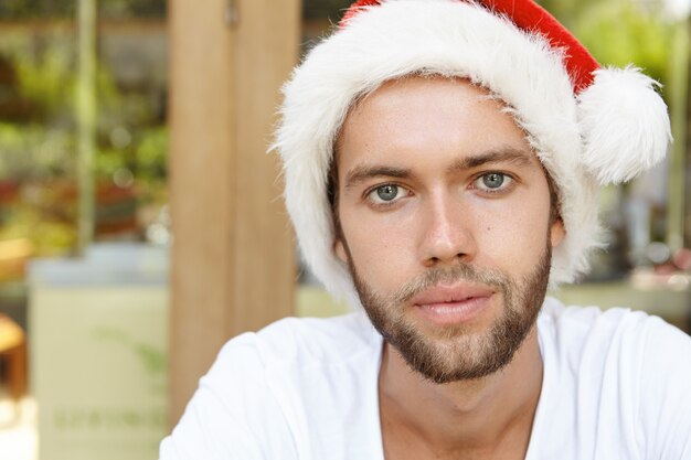 Close up portrait of attractive young Caucasian man wearing casual t-shirt and Santa Claus hat looking at camera with confident smile, spending nice time at cafe