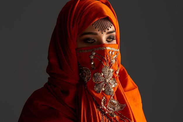 Free photo close-up portrait of an attractive young female with beautiful smoky eyes and jewelry on the forehead, wearing the terracotta hijab decorated with sequins. she is posing and looking down on a dark bac