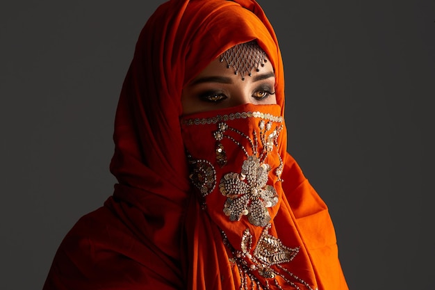 Close-up portrait of an attractive young female with beautiful smoky eyes and jewelry on the forehead, wearing the terracotta hijab decorated with sequins. She is posing and looking down on a dark bac