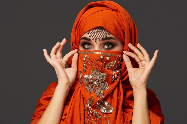 Close-up portrait of an attractive young woman with beautiful smoky eyes and fine jewelry on the forehead, wearing the terracotta hijab decorated with sequins. She is holding the shawl with her hands