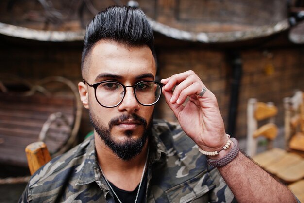 Close up portrait of awesome beautiful tall ararbian beard macho man in glasses and military jacket posed outdoor sitting on chair against wooden house