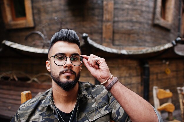 Close up portrait of awesome beautiful tall ararbian beard macho man in glasses and military jacket posed outdoor sitting on chair against wooden house