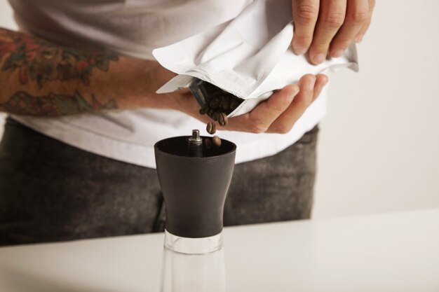 Close up portrait of a barista in white t-shirt and jeans pouring coffee beans into a modern small burr grinder