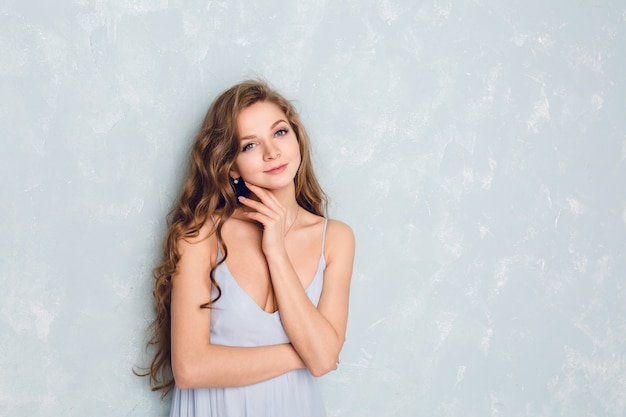 Close-up portrait of a beautiful blond girl with curly hair standing in a studio.  She holds her hand near chin