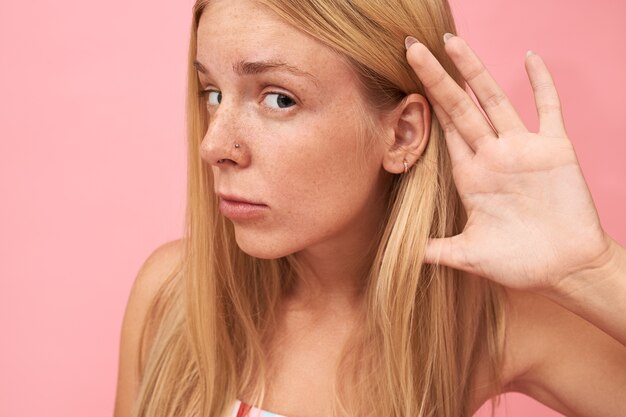 Close up Portrait of beautiful blonde teenage girl with long loose hair, freckles and nose piercing placing hand at her ear, having curious nosy look