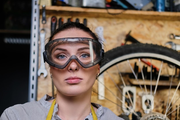 Free photo close-up portrait of a beautiful brunette female wearing working clothes, apron and goggles, standing in a workshop against wall tools.