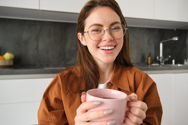 眼鏡をかぶった美しい茶色の女性のクローズアップポートレート ⁇ カップ茶でキッチンに座って休息しています ⁇ 