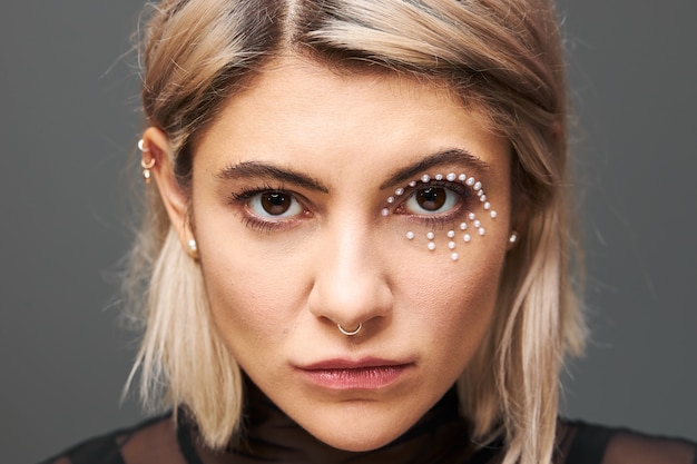 Close up portrait of beautiful charming young blonde female model with short dyed hair staring right, having mysterious intent gaze, wearing black dress, face piercing and glamorous make up