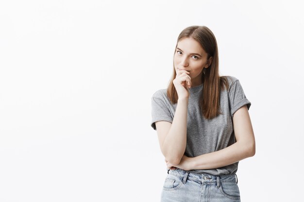 Close up portrait of beautiful dark-haired caucasian young woman with long hair in grey t-shirt and jeans with thoughtful face expression, smiling, holding hand near mouth,