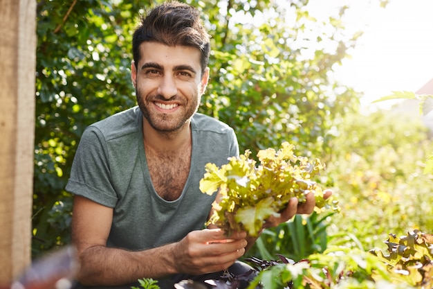 Крупным планом портрет красивого темнокожего бородатого кавказского фермера, улыбающегося, работающего в саду, собирающего листья салата, готовящегося к вечерней встрече с друзьями в своем доме