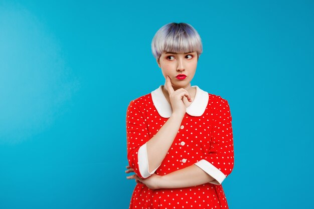 Close up portrait beautiful dollish girl with short light violet hair wearing red dress over blue wall