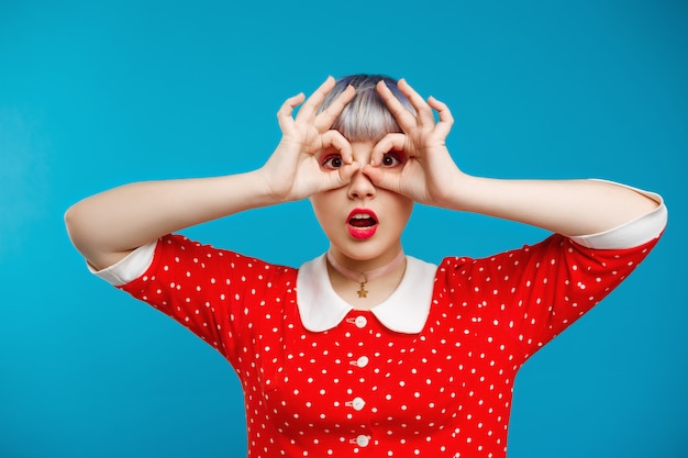 Close up portrait beautiful dollish girl with short light violet hair wearing red dress over blue wall