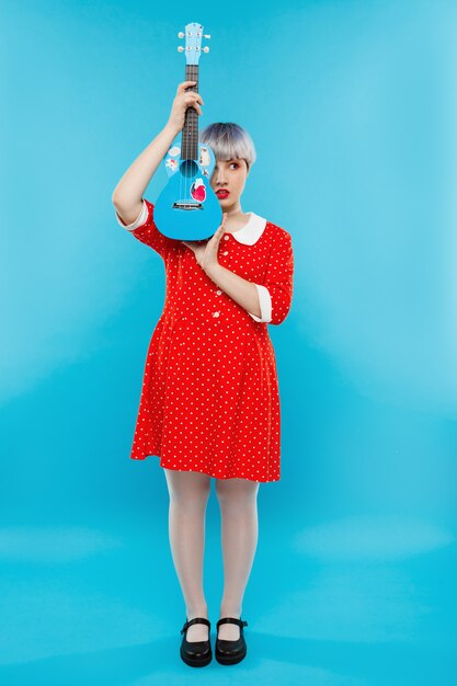 Close up portrait of beautiful dollish girl with short light violet hair wearing red dress holding ukulele over blue wall